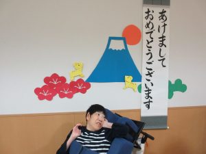 白い壁に富士山の切り絵とあけましておめでとうございますの文字をバックに男性利用者さんが座っている様子