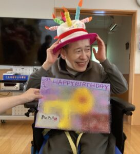 ローソクの飾りのついたお誕生日帽子を利用者さんが被ってお誕生日カードと一緒微笑む利用者さんの写真